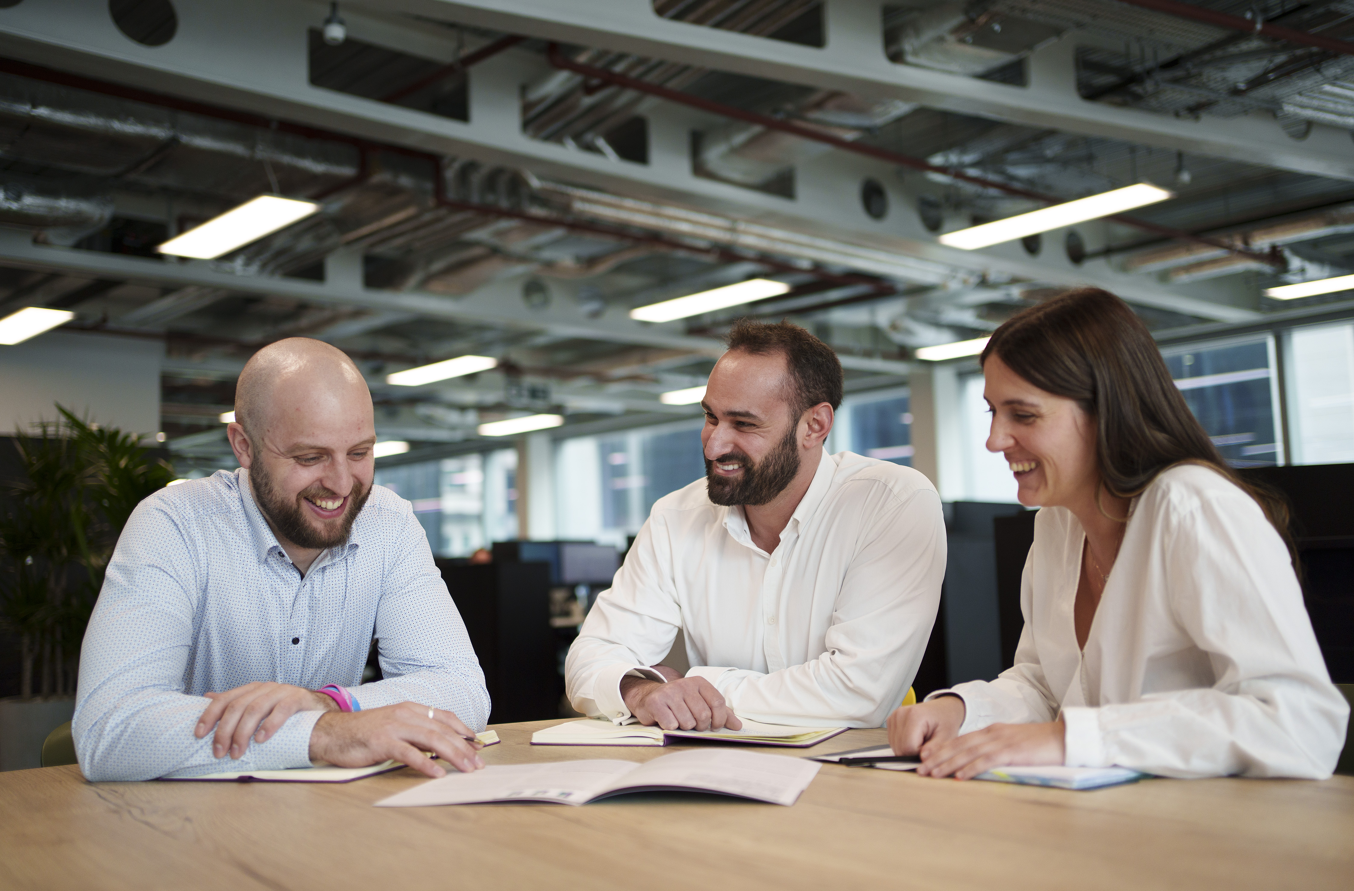 Three employees gathered round an informal meeting table with open plan office in background